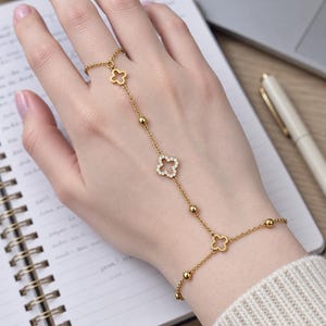 Peut inclure: Une chaîne de main dorée au design délicat. Le bijou se compose de trois breloques en forme de trèfle, dont l'une est incrustée de petites pierres claires, et de plusieurs perles dorées espacées le long de la chaîne.