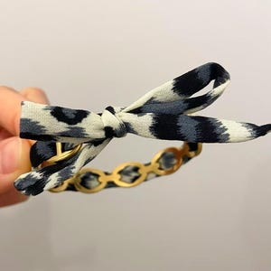 Può includere: Un bracciale caratterizzato da una catena in metallo dorato con maglie circolari, parzialmente avvolta in un nastro di tessuto con motivo animalier nero, grigio e bianco, annodato in un fiocco decorativo sulla parte superiore del bracciale.