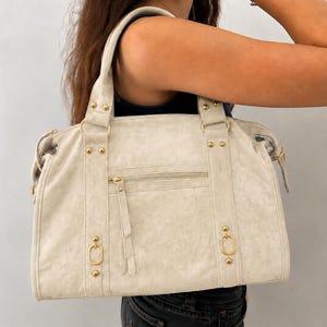 May include: A cream-colored handbag with a distressed texture, featuring gold-tone metal hardware, including studs, rings, and a zippered front pocket. The bag has two top handles and a structured rectangular shape with vertical stitched detailing.
