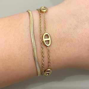Peut inclure: Un ensemble de deux bracelets dorés. L'un est une chaîne à chevrons plate et flexible, et l'autre est une fine chaîne à maillons comportant trois éléments ovales métalliques régulièrement espacés avec une barre horizontale au centre.