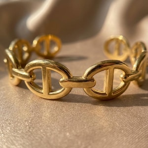 Op de afbeelding: Een goudkleurige armband met een schakelontwerp. De ovale schakels hebben een centrale staaf. De armband is gemaakt van glanzend, metaalachtig materiaal. Het ontwerp is elegant.