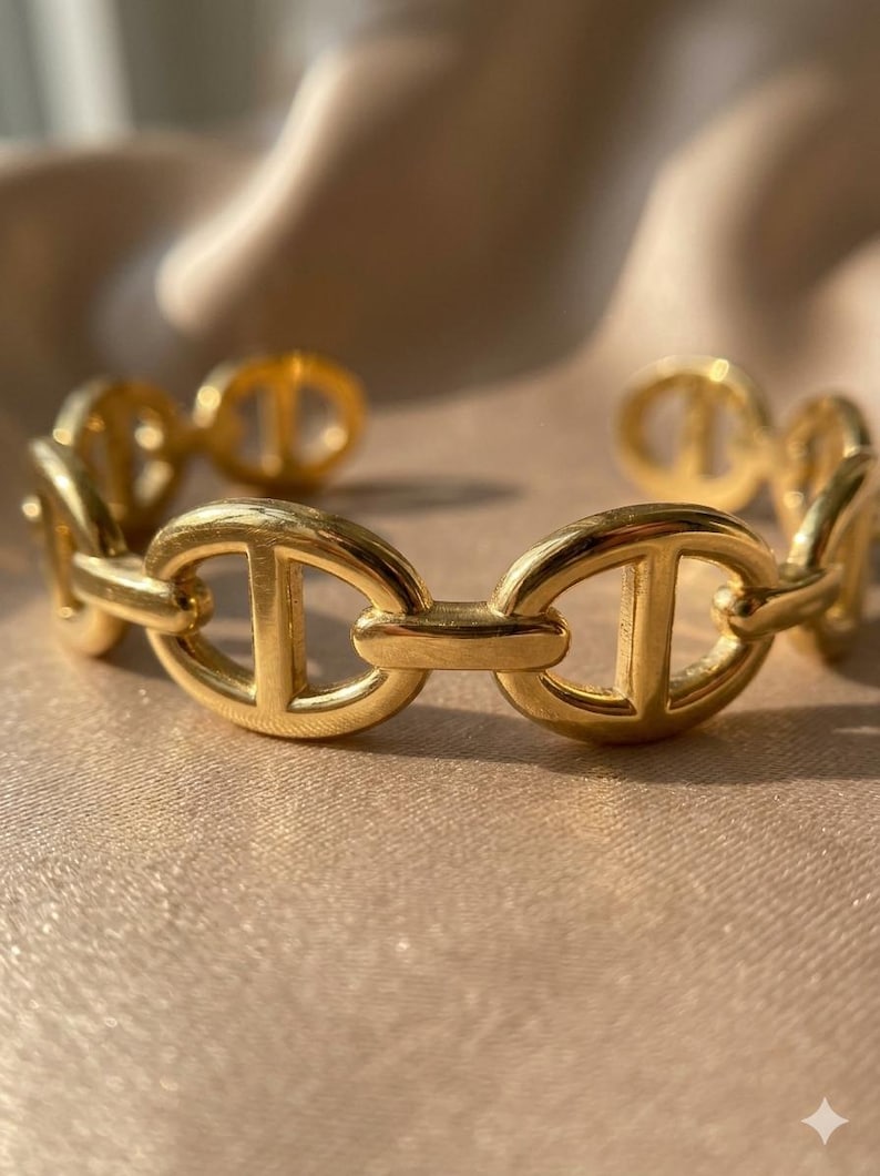 Op de afbeelding: Een goudkleurige armband met een schakelontwerp. De ovale schakels hebben een centrale staaf. De armband is gemaakt van glanzend, metaalachtig materiaal. Het ontwerp is elegant.