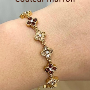 Op de afbeelding: Een goudkleurige kettingarmband met een reeks klaverbladvormige bedels. Elke bedel is bezet met kleine, sprankelende bruine en heldere kristallen. De tekst "Couleur marron" is zichtbaar bovenaan de afbeelding en geeft de bruine kleur aan.