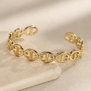 Peut inclure: Un bracelet manchette doré composé d'une série de maillons circulaires répétitifs, chacun doté d'une barre horizontale centrale. Ce modèle ouvert est réalisé en métal poli, formant une structure en chaîne continue autour du poignet.