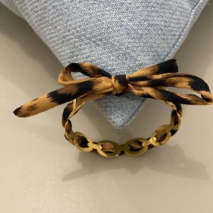 Peut inclure: Un bracelet doré avec un motif de maillons ovales, orné d'un nœud en tissu à imprimé léopard. Le bracelet est posé sur un fond de tissu bleu clair, créant un contraste de textures et de couleurs.