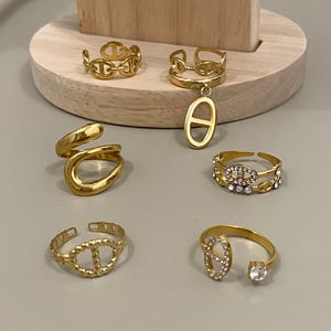 Op de afbeelding: Een collectie van zes goudkleurige ringen met diverse ontwerpen, waaronder schakelpatronen, een bungelende ovale bedel, een gladde wikkelstijl en ringen versierd met heldere kristallen, gepresenteerd op een lichtgekleurd oppervlak.