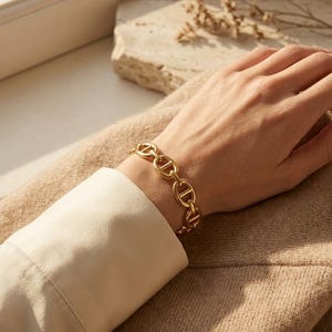 Op de afbeelding: Een goudkleurige kettingarmband met een reeks in elkaar grijpende ovale schakels. De armband wordt om een pols gedragen, met een cr&egrave;mekleurig shirt met lange mouwen zichtbaar. De achtergrond omvat een neutraal getinte trui en een steen.
