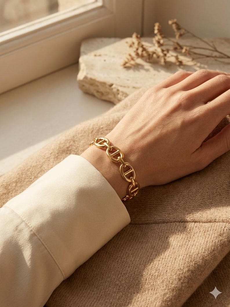 Op de afbeelding: Een goudkleurige kettingarmband met een reeks in elkaar grijpende ovale schakels. De armband wordt om een pols gedragen, met een cr&egrave;mekleurig shirt met lange mouwen zichtbaar. De achtergrond omvat een neutraal getinte trui en een steen.