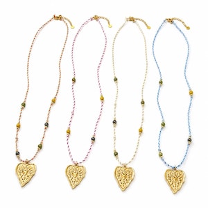 Peut inclure: Quatre colliers avec des cordons tressés orange, rose, blanc et bleu, ornés de petites perles vertes et sombres. Chaque collier comporte un pendentif doré en forme de cœur avec un motif soleil en relief et un petit cœur central.