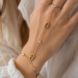Peut inclure: Un bracelet de main doré composé d'une fine chaîne reliant une bague à un bracelet de poignet. Le bijou est orné de petits cristaux ronds transparents et de deux maillons dorés de forme ovale avec une barre horizontale au centre.