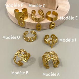 Pode incluir: Uma coleção de sete anéis de metal dourado com vários designs de elos e bandas. Os anéis estão rotulados como Modèle F, C, E, O, I, B e A. Alguns anéis apresentam pingentes pendentes, enquanto outros exibem padrões de elos entrelaçados.