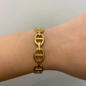 Op de afbeelding: Een goudkleurige armband met een schakelkettingontwerp. De armband is gemaakt van metaal en heeft een reeks ovale schakels die zijn verbonden door horizontale staven. De armband wordt om de pols gedragen.
