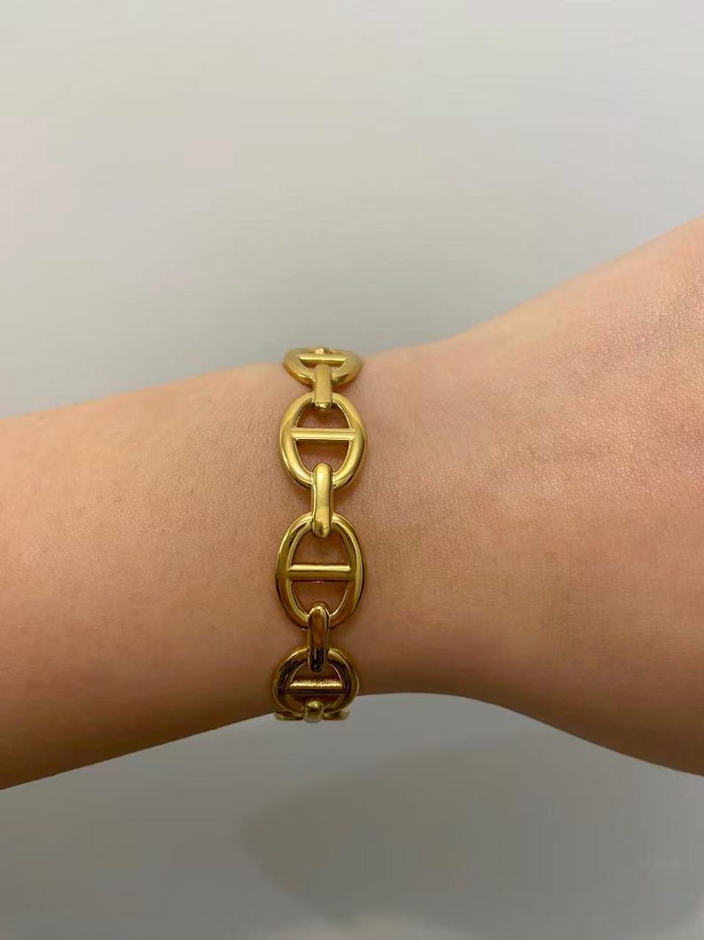 Op de afbeelding: Een goudkleurige armband met een schakelkettingontwerp. De armband is gemaakt van metaal en heeft een reeks ovale schakels die zijn verbonden door horizontale staven. De armband wordt om de pols gedragen.