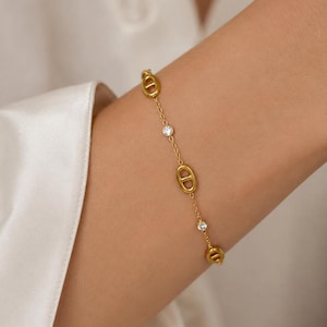 Peut inclure: Un bracelet doré orné de maillons ovales et de petites pierres claires scintillantes. Le bracelet est présenté sur un bras, sur un fond neutre et doux. Le bijou est délicat et élégant.