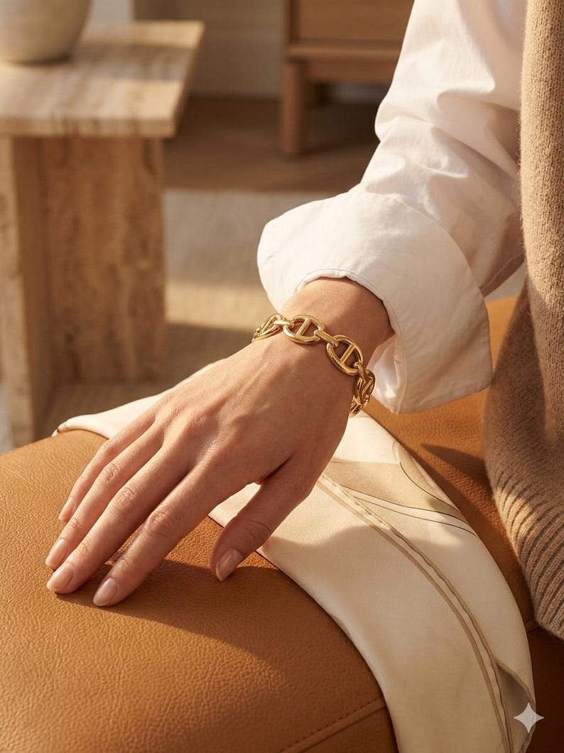 Op de afbeelding: Een goudkleurige schakelarmband wordt om een pols gedragen. De armband is gemaakt van in elkaar grijpende ovale en D-vormige schakels. De persoon draagt een wit overhemd met lange mouwen. De achtergrond omvat een bruin leren oppervlak en een beige sjaal.