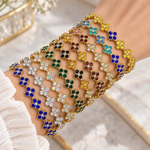 Peut inclure: Une collection de plusieurs bracelets en métal doré présentant un motif répétitif de trèfle à quatre feuilles. Chaque trèfle est orné de petits cristaux vibrants et multicolores, notamment des pierres bleues, vertes, jaunes, claires et rouge foncé.