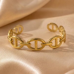 Op de afbeelding: Een goudkleurige manchetarmband met een schakelkettingontwerp. De schakels zijn ovaal van vorm met een verticale balk in het midden. De armband is aan de uiteinden open en ligt op een satijnen oppervlak.