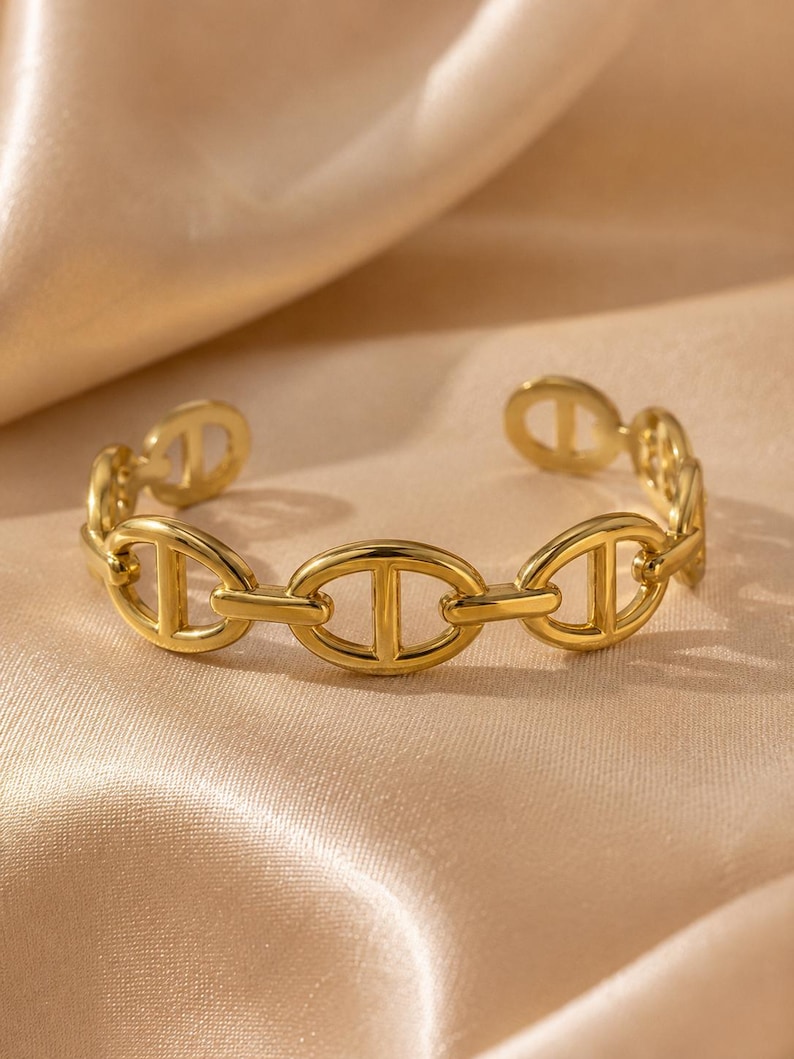 Op de afbeelding: Een goudkleurige manchetarmband met een schakelkettingontwerp. De schakels zijn ovaal van vorm met een verticale balk in het midden. De armband is aan de uiteinden open en ligt op een satijnen oppervlak.