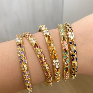 Peut inclure: Cinq bracelets joncs articulés en métal doré avec un motif floral ajouré rempli d'émail coloré dans des tons violet, rose, orange, vert et bleu, agrémentés de petits cristaux transparents incrustés au centre des fleurs.
