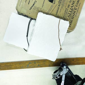 May include: Two white, textured paper notebooks with black and brown string closures. An antique camera and a wooden ruler are also visible. The notebooks are next to a vintage container with text.