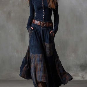 Puede incluir: Un vestido azul marino de manga larga con cuello alto y cierre de botones. El vestido presenta un cinturón de cuero marrón y una falda larga con parches a cuadros. Botas marrones completan el atuendo, creando un estilo bohemio.