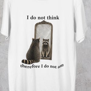 Puede incluir: Camiseta blanca con un diseño de mapache. El mapache se mira en un espejo. El espejo está enmarcado y el texto "I do not think therefore I do not am" está impreso encima y debajo del espejo.