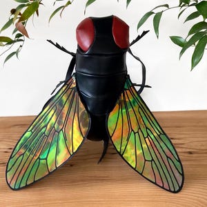 May include: A distinctive, black fly-shaped bag with iridescent wings. The bag has large, red mesh eyes and detailed wing patterns in green, yellow, and orange. The bag is displayed on a wooden surface.