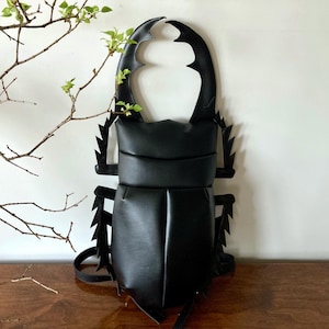 Pode incluir: Uma mochila preta em forma de besouro veado, com chifres grandes e curvos e detalhes de pernas pontiagudas. A bolsa tem um corpo arredondado e alças ajustáveis. Um acessório único e atraente.