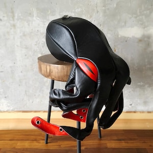 May include: Black leather octopus-shaped backpack with red detailing. The backpack features a rounded top, a zip, and multiple tentacle-like straps. Red accents are visible on the sides and tentacles. The backpack is on a wooden stool.