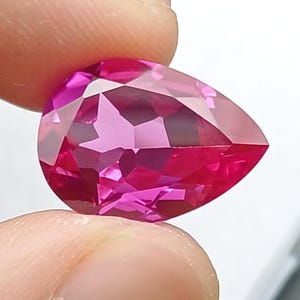 Op de afbeelding: Een gefacetteerde, peervormige edelsteen in een levendige roze tint. De edelsteen wordt tussen twee vingers gehouden, waarbij de gepolijste oppervlakken en interne reflecties worden getoond. De achtergrond is een witte doos en een donkergrijze ondergrond.