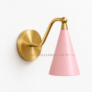 Könnte beinhalten: Eine Wandleuchte mit einer runden Montageplatte aus gebürstetem Messing, einem geschwungenen Messingarm und einem konischen Lampenschirm in zartem Rosa. Der Text BRASS BRILLIANCE GIFTS ist horizontal mittig auf der Leuchte platziert.