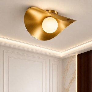 Könnte beinhalten: Eine moderne Deckenleuchte mit goldener Oberfläche, die ein geschwungenes, blattartiges Design aufweist. Die Leuchte hat eine kugelförmige Glühbirne aus Milchglas in der Mitte. Die Buchstaben "BBG" sind am Rand der Leuchte sichtbar.