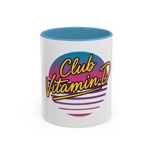 Op de afbeelding: Witte keramische mok met een blauwe binnenkant. De mok heeft de woorden "Club Vitamin D" in gele letters over een retro zonsondergangontwerp. Het ontwerp heeft een verloop van paars, roze en blauw met horizontale paarse lijnen.