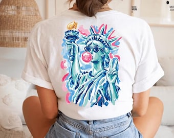May include: White t-shirt featuring a colorful illustration of the Statue of Liberty wearing sunglasses and blowing a bubblegum bubble. The design includes blue, pink, and yellow accents. The shirt has short sleeves.