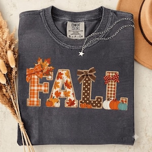 Könnte beinhalten: Ein anthrazitfarbenes T-Shirt mit dem Wort FALL in großen Blockbuchstaben. Jeder Buchstabe hat ein einzigartiges Muster, darunter orangefarbenes Karomuster, Herbstblätter, braune Punkte und braunes Karomuster, verziert mit Schleifen und Kürbissen.