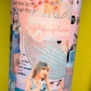 May include: A cylindrical tumbler featuring a collage of pink and blue imagery, including photographs of a performer in a sequined bodysuit, song titles like Paper Rings and Cornelia Street, and the text Meet me in the afterglow in cursive script.