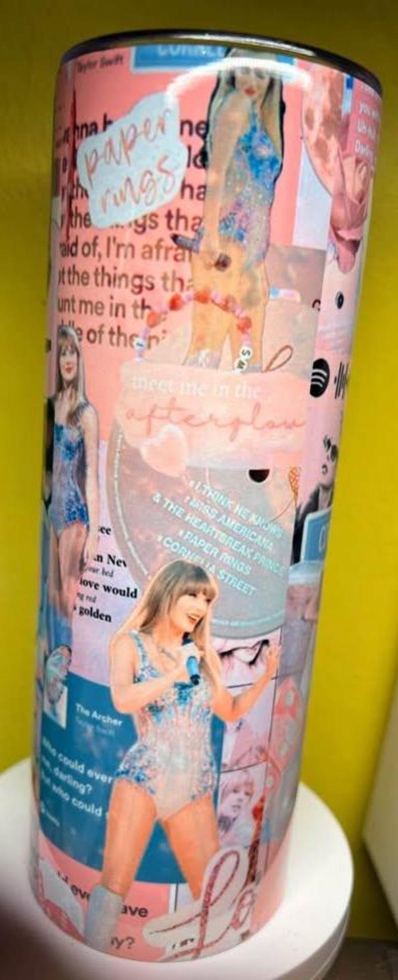 May include: A cylindrical tumbler featuring a collage of pink and blue imagery, including photographs of a performer in a sequined bodysuit, song titles like Paper Rings and Cornelia Street, and the text Meet me in the afterglow in cursive script.