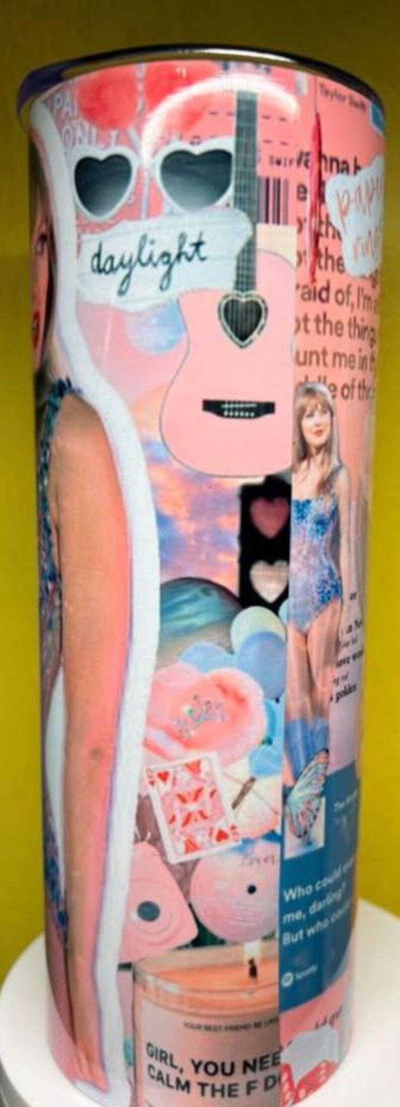 May include: A cylindrical tumbler featuring a collage of pink and blue imagery, including a guitar, heart-shaped sunglasses, a playing card, and text such as daylight, paper rings, and girl, you need to calm the f down, with photographic portraits.