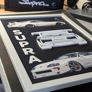 Puede incluir: Una pieza de arte mural enmarcada que presenta un coche Toyota Supra blanco. La obra incluye una vista de perfil, una vista frontal y un recorte blanco en relieve 3D de la parte delantera del coche. La palabra SUPRA está impresa verticalmente.