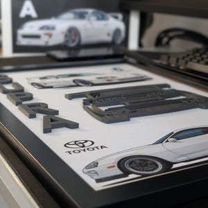 May include: A framed artwork featuring a white Toyota Supra car design. The word "SUPRA" is in gray 3D letters. The Toyota logo is visible. The artwork is in a black frame.