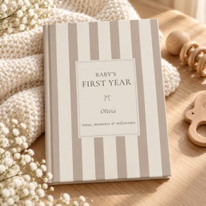 Puede incluir: Un libro de recuerdos de tapa dura con rayas verticales beige y blancas. La portada presenta un rectángulo blanco central con el texto BABY'S FIRST YEAR, un pequeño icono de lazo, el nombre Olivia y las palabras notes, moments & milestones.