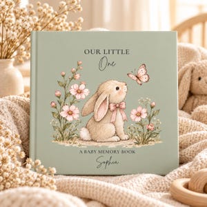 May include: A sage green baby memory book featuring a central illustration of a beige bunny with a pink bow, surrounded by pink flowers and a butterfly. The cover text reads: OUR LITTLE One A BABY MEMORY BOOK Sophia.