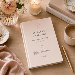 May include: A light pink hardcover journal featuring the printed text "IT TAKES A VILLAGE thank you for being part of ours" with two small pink hearts, the name "Mrs. Anderson" in script, and the year "2026" centered on the front cover.