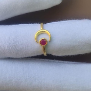 Puede incluir: Un anillo de oro con forma de media luna situada sobre una pequeña piedra preciosa redonda de color rojo vibrante. La banda es fina y pulida, mostrando un tono oro amarillo brillante que contrasta con el color rojo intenso de la piedra.