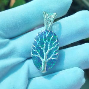 Könnte beinhalten: Ein ovaler Anhänger mit einem dunkelblau und grün schimmernden Stein, der mit silberfarbenem Draht umwickelt ist. Der Draht ist kunstvoll zu einem Baum geformt, dessen Äste sich über die Oberfläche des Steins erstrecken, mit einer Öse oben.