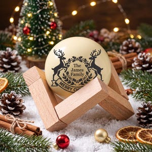 Peut inclure: Une boule de Noël sphérique de couleur crème reposant sur un support en bois clair. La boule présente un motif noir composé de deux rennes entourant une couronne centrale avec le texte "The James Family" et "MERRY CHRISTMAS" en dessous.