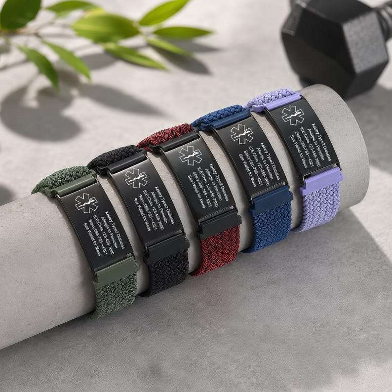 May include: Five medical alert bracelets with braided nylon bands in olive green, black, maroon, navy blue, and lavender. Each features a black metal plate engraved with a Star of Life symbol and text: Kelsey, Type 2 Diabetes, Allergic to Penicillin.