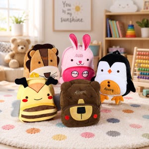 Puede incluir: Cinco mochilas de peluche con forma de animal que representan un león, un conejo rosa, un pingüino, una abeja y un oso. Cada mochila muestra un nombre bordado en la parte delantera: Daniel, Mille, Avery, Lucilla y Alvaro. Tienen texturas suaves.