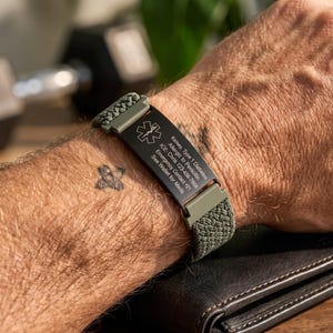 May include: An olive green woven medical alert bracelet featuring a black metal plate with the Star of Life symbol and engraved text: Kelsey, Type 1 Diabetes, Allergic to Penicillin, ICE: Chris 123-456-7886, Emergency Contact +21, See Wallet for Meds.