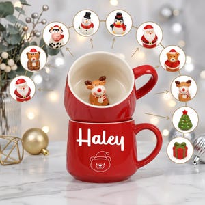 Könnte beinhalten: Ein roter Keramikbecher mit weißem Innenraum und einer 3D-Rentierfigur im Inneren. Außen ist der Name Haley in weißer Schrift über einem weißen Strichmännchen-Bären mit Weihnachtsmütze abgebildet. Um den Becher herum sind kreisförmige Symbole von Festtagsfiguren.